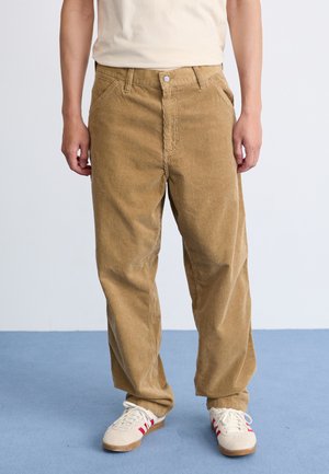 Persona de pie con pantalones de pana beige y zapatillas cremadas con rayas rojas, sobre un suelo azul y un fondo de pared blanca.