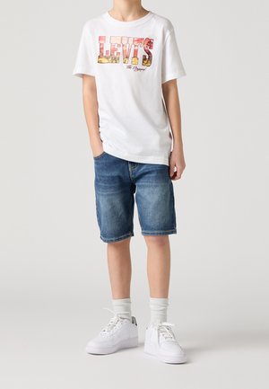 Enfant portant un t-shirt blanc Levi's, un short en jean, des chaussettes blanches et des baskets blanches, debout devant un fond uni.