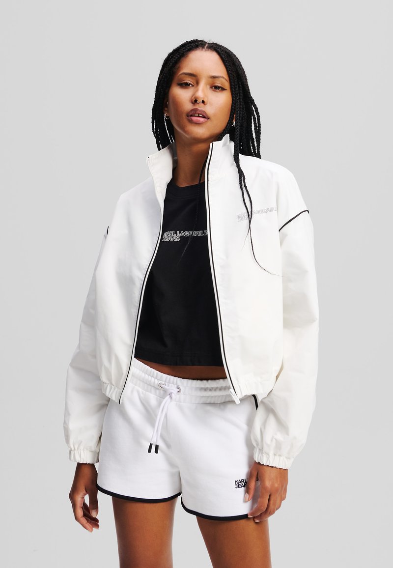 Witte cropped jasje met een hoge kraag, zwarte accenten en elastische manchetten, gedragen over een zwart T-shirt en witte shorts met een trekkoord.