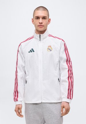 Muž oblečený v bielej bunde na zips s červenými pruhmi na rukávoch a logami Real Madrid a Adidas, v sivej teplákovej súprave.