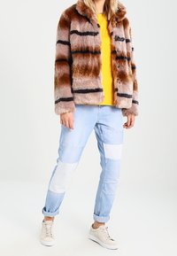 Kurtka z futerka syntetycznego w odcieniach brązu z czarnymi paskami, zestawiona z żółtą bluzką i jasnoniebieskimi dżinsami z naszywkami; cechuje się luźnym krojem.