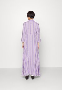 Lång, lila klänning med vitt zigzag-mönster, med hög krage och flödande ärmar. Tyget verkar lätt och texturerat.