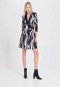 Schwarz-weißes Kleid mit gewelltem Muster, doppelt geknöpft, Dreiviertelärmel und einer taillierten Passform. Kombiniert mit schwarzen Stiefeletten.