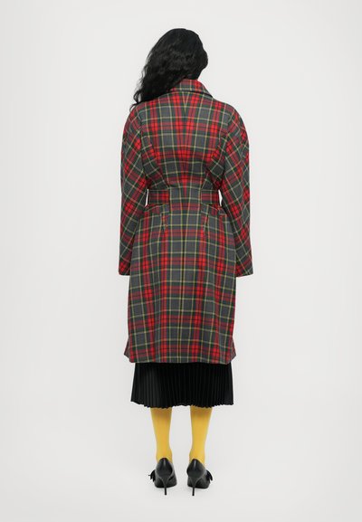 Cappotto lungo a scacchi rosso, nero e giallo con vita regolabile, cuciture in vista e ampie revers, abbinato a una gonna nera plissettata e collant gialli.