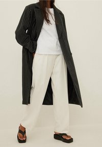 Cappotto lungo a righe nere, t-shirt bianca e pantaloni bianchi a gamba larga. Sandali con zeppa neri accentuano l'aspetto moderno dell'outfit.