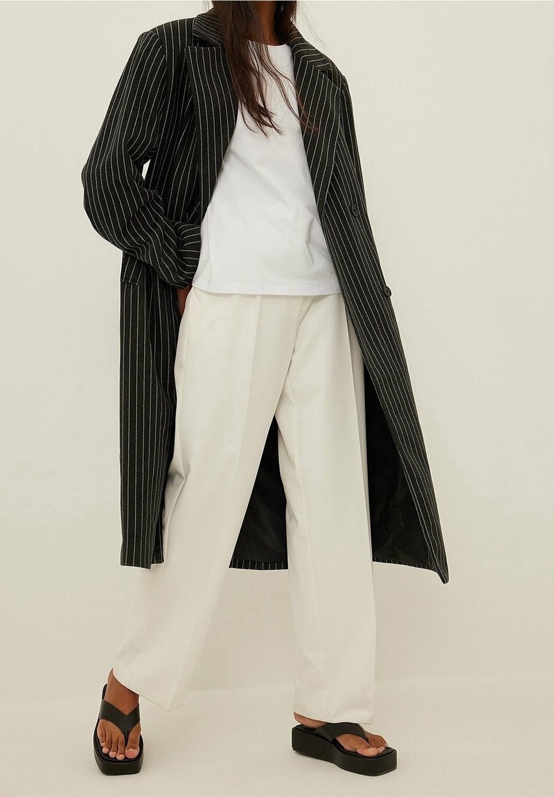Cappotto lungo a righe nere, t-shirt bianca e pantaloni bianchi a gamba larga. Sandali con zeppa neri accentuano l'aspetto moderno dell'outfit.
