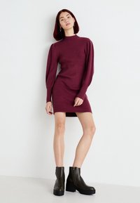 Vestido de punto burdeos con textura acanalada, cuello alto y mangas abullonadas. Combinado con botas de color verde oscuro de longitud a la altura del tobillo con tacón cuadrado.