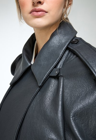 Close-up van een persoon die een zwarte leren jas draagt met een brede kraag en een schouderriem met knoop, lippen en kin zichtbaar, neutrale achtergrond.
