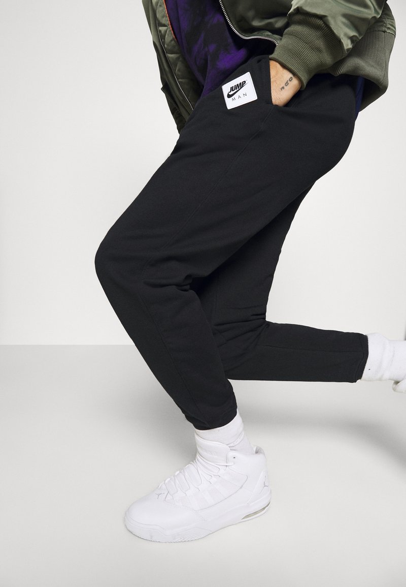 Black sweatpants with pockets, drawstring waist, and a white sports label. Worn with white athletic shoes and visible white socks.