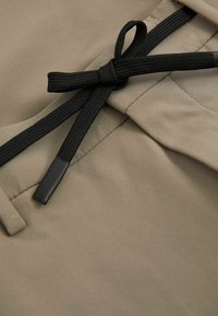 Material bej cu o textură netedă, având un șnur negru legat sub formă de fundiță, accentuat de un vârf din plastic, poziționat pe o secțiune pliată.