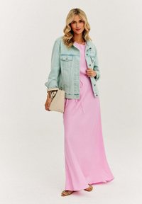 Jasnoniebieska dżinsowa kurtka na różowej sukience maxi. Sukienka ma elastyczny pas, a modelka trzyma beżową torbę i nosi buty w wzory.