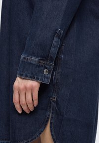 Tmavomodré džínsové šaty-košeľa s bodovým golierom, zapínaným manžetou a bočnými rozparky. Má viditeľné stehy a tkanú značkovú štítok.