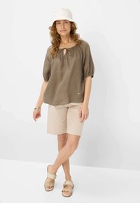 Blusa di lino marrone con maniche a sbuffo e dettaglio con fiocco, abbinata a pantaloni corti beige e sandali beige chiari. Modello che indossa un cappello a secchiello bianco.
