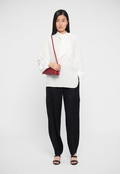 White blouse with bow tie detail, black cargo pants with side pockets, red handbag, black strappy sandals, model stands against a plain background.