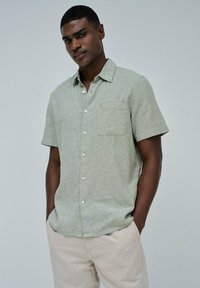Camisa de manga corta verde claro en tejido texturizado con un bolsillo de parche, botones en el frente y cuello con punta. Se lleva con pantalones beige claros.