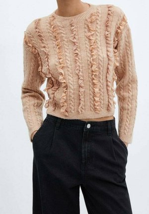 Femme portant un pull beige en maille avec des volants verticaux, associé à un pantalon noir taille haute, mains dans les poches, sur fond uni.