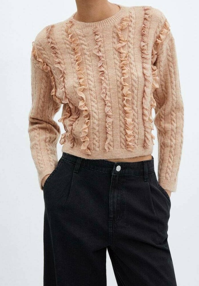 Femme portant un pull beige en maille avec des volants verticaux, associé à un pantalon noir taille haute, mains dans les poches, sur fond uni.