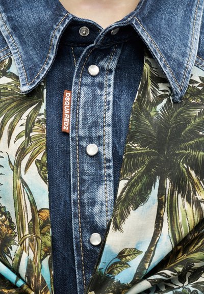Πουκάμισο από τζιν με λευκά κουμπιά πατιλέτας, καφέ ετικέτα "DSQUARED2" και τροπικό ύφασμα με σχέδιο φοίνικες τοποθετημένο πάνω του.