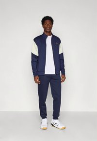 Marineblå atletisk treningsdress med glidelåsjakke med lysegrønne detaljer og matchende bukser. Kombinert med hvite treningssko.