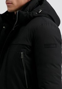 Zwarte pufferjack met een hoge kraag en capuchon, voorzien van een zijzak met rits, horizontale naaddetails en een logopatch op de mouw.