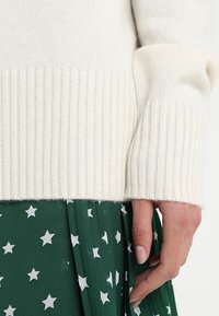 Kremowy sweter z prążkowanym dołem i mankietami, noszony na zielonej spódnicy plisowanej z białymi wzorami w gwiazdki. Miękka tekstura, prosty design i warstwowy wygląd.