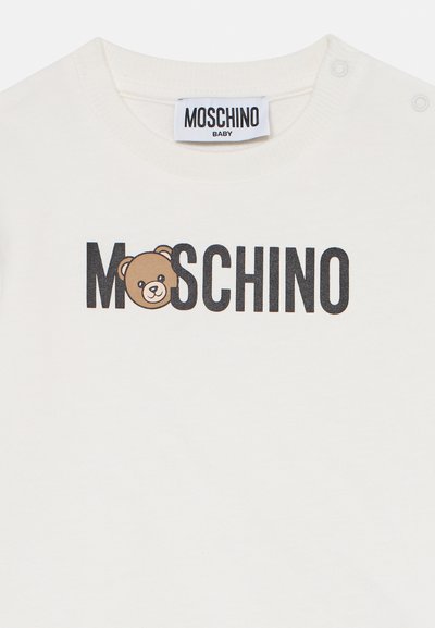 Vit babyskorta med svart "MOSCHINO"-text där en leende teddy-björn ansikte ersätter bokstaven "O" i logotypen.