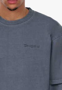 Niebieski, oversize'owy sweter wykonany z miękkiego materiału, z ryflowanym dekoltem i haftowanym logo "Dropsize" w ciemnym kolorze na klatce piersiowej.
