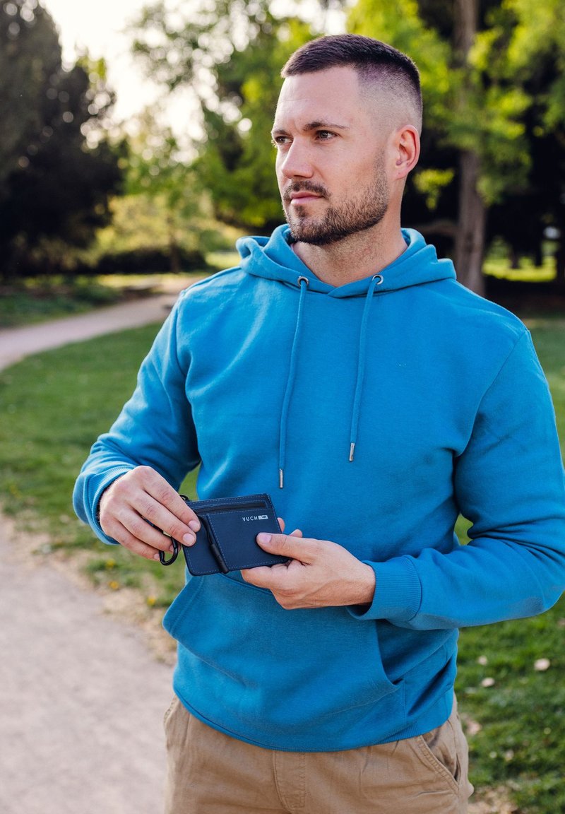 Blue cotton hoodie with drawstrings, worn by a person holding a dark blue cardholder with a logo. Background shows a grassy park path.