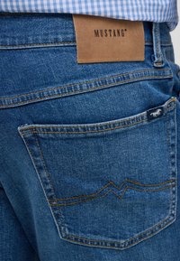 Niebieskie dżinsy z kontrastującymi pomarańczowymi przeszyciami, z skórzaną metką z napisem "MUSTANG" oraz charakterystycznym wzorem kieszeni.