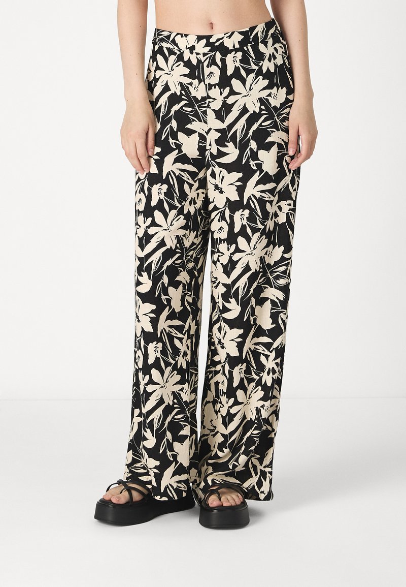 Wide-leg trousers in black fabric featuring a floral pattern in cream. Smooth texture with a high waist and side pockets. Paired with black sandals.