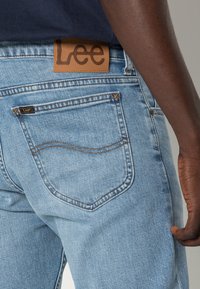 Světle modré džíny z denimu s koženou nášivkou značky Lee na pase, klasický pětikapsový design a kontrastní švy.