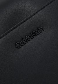 Borsa a tracolla in pelle nera testurizzata con logo "Calvin Klein" impresso. Dettagli cuciti che mettono in risalto il design.