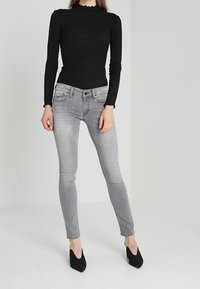 Top negro de manga larga con canalé y escote con volantes, combinado con unos pantalones pitillo gris claro y tacones negros puntiagudos, sobre un fondo neutro.
