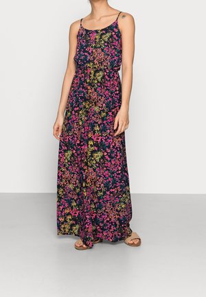 Woman wearing a sleeveless, floral-print maxi dress with thin straps and flat sandals, standing against a plain light background.
