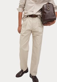 Beiga plisserade byxor med slim fit, tillsammans med en ljusbeige långärmad skjorta och mörkbruna loafers; hållande en mörkbrun väska.