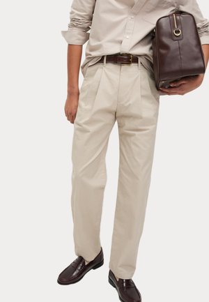 Pantalones plisados beige de corte ajustado, combinados con una camisa de manga larga beige claro y unos mocasines marrón oscuro; sosteniendo un bolso marrón oscuro.