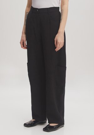 Zwarte wijde broek met twee zijzakken, gemaakt van een zachte stof. De broek heeft een hoge taille en een klassieke pasvorm, gecombineerd met zwarte platte schoenen.
