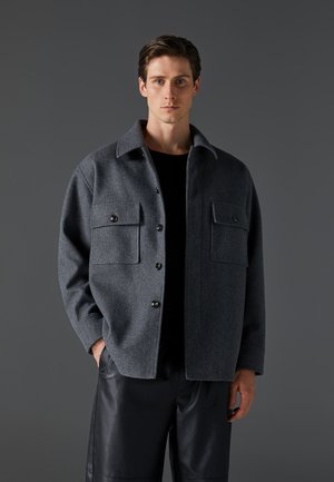 Szürke gyapjú keverék ingdzseki, gallérral, két elülső zsebbel, sötét gombokkal és laza fazonnal, fekete bőrnadrággal párosítva.