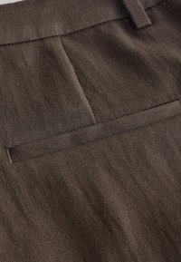 Gros plan sur un tissu marron montrant une poche passepoilée horizontale avec des coutures visibles et des détails de texture.