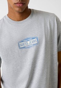 Szara bawełniana koszulka z niebiesko-białym logo "GUESS JEANS" na piersi, z okrągłym dekoltem i krótkimi rękawami. Gładka faktura.