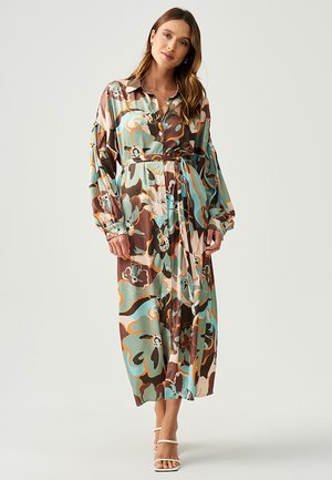 Long, button-up dress in shiny fabric featuring a colorful floral pattern with brown, teal, and peach tones. Belts at the waist, long sleeves.