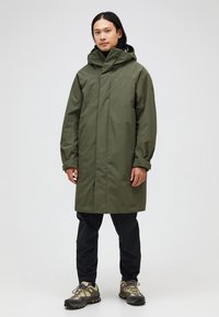 Olive green waterproof coat with a hood, front buttons, long sleeves, and a straight cut, paired with black pants and hiking shoes.