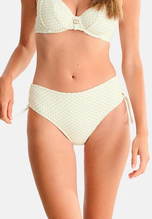 Femme ajustant un bas de bikini taille haute texturé crème avec des liens latéraux, portant un haut de soutien-gorge assorti, sur fond uni.