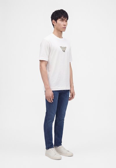 Νεαρός άνδρας που φοράει λευκό t-shirt με γραφικό λογότυπο, στενά μπλε τζιν και λευκά αθλητικά παπούτσια, μπροστά σε απλό φόντο.