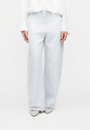 Personne portant un pantalon large gris clair, un haut blanc à manches longues et des chaussures blanches pointues, debout devant un fond uni.