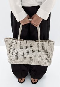 Geweven handtas in lichtbeige met een gestructureerd patroon, voorzien van twee handvatten en een ruime open bovenkant. Gecombineerd met zwarte wijde broek.