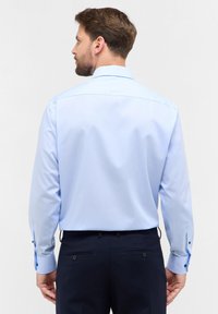 Camicia azzurra chiaro a maniche lunghe con colletto button-down, texture liscia e dettagli dei bottoni a contrasto sui polsini. Indossata con pantaloni scuri.