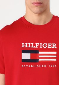 Červené bavlněné tričko s velkým vyšívaným vzorem vlajky v námořnické modři, bílé a červené barvě, s textem "HILFIGER" a "ESTABLISHED 1985" v bílé barvě.