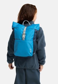 Mochila azul con un diseño de rollo en la parte superior, correa gris ajustable y bolsillos laterales de malla. Logo visible en la parte frontal. Tela texturizada.