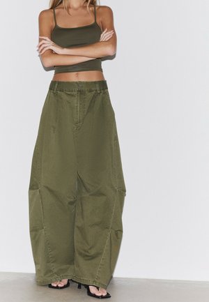Femme portant un pantalon large vert olive, un débardeur court assorti et des sandales à talons noires, debout, bras croisés, sur fond blanc.